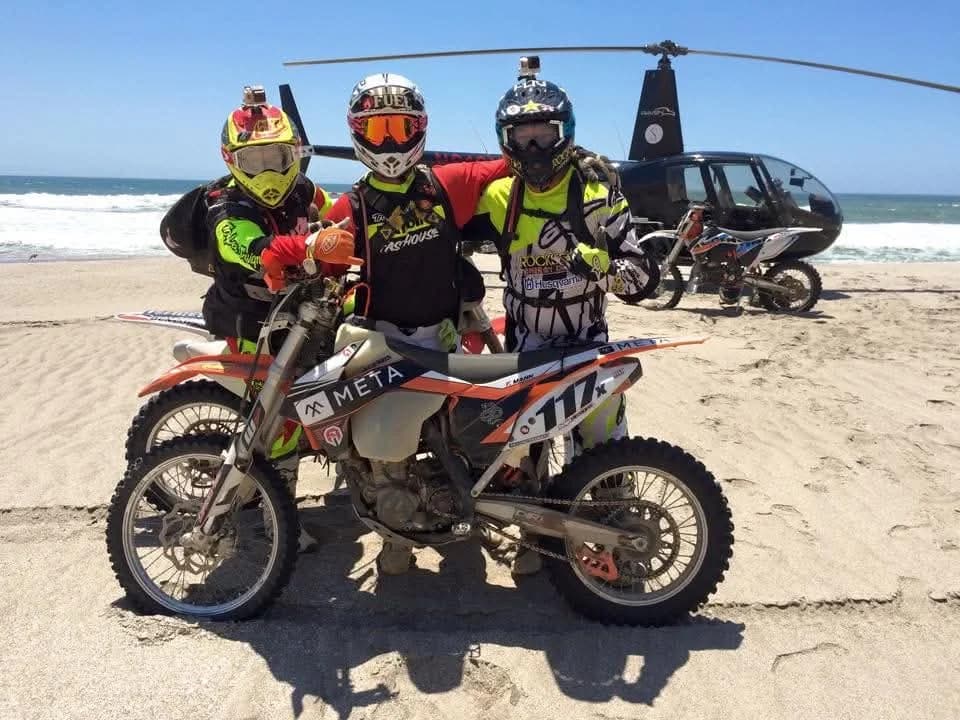 Three riders on dirt bikes on a beach, a helicopter behind them