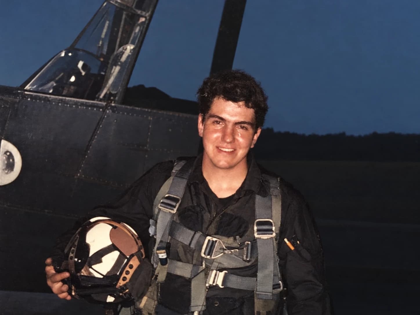 John Mann Sr. as a young man in flight gear, holding his helmet beside an aircraft